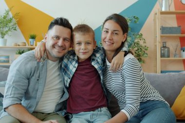 portrait of happy family mother, father and son smiling looking at camera sitting on couch in modern apartment. People and lifestyle concept.